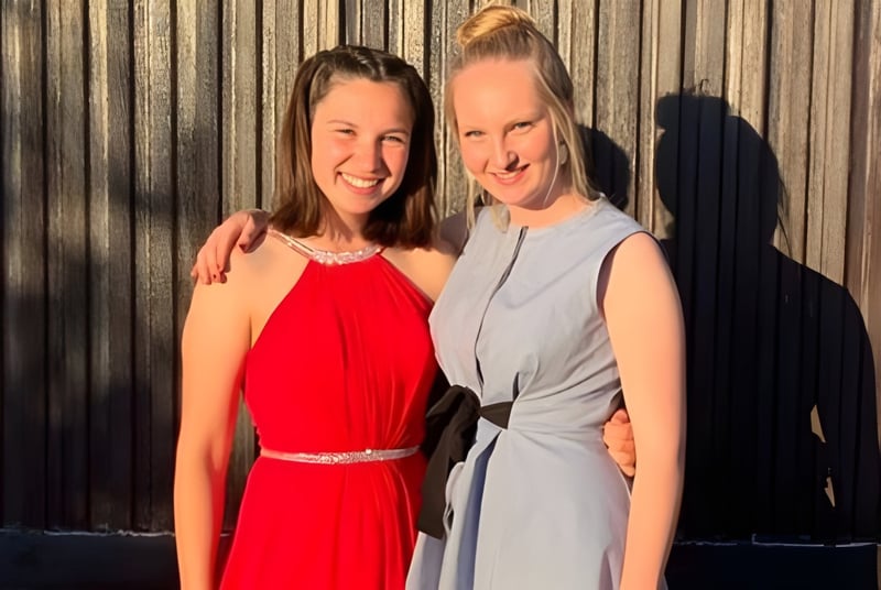 Zwei lächelnde Frauen stehen vor einem Holzzaun auf dem Gelände der Stonar School.