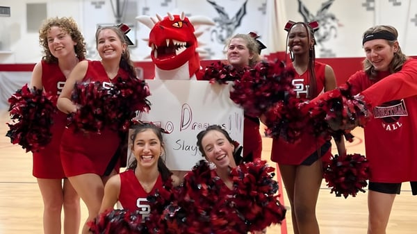 Eine Gruppe Cheerleader der Stuart Hall School posiert in roten und schwarzen Uniformen mit Pompons und einer großen roten Maskottchenkostüm in einer Turnhalle.
