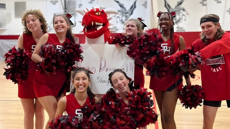 Eine Gruppe Cheerleader der Stuart Hall School posiert in roten und schwarzen Uniformen mit Pompons und einer großen roten Maskottchenkostüm in einer Turnhalle.