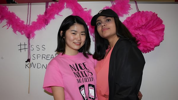 Zwei Schülerinnen der Summerland Secondary School posieren vor einer Wand mit großen rosa Federdekorationen.