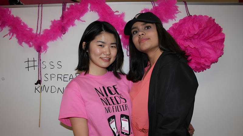 Zwei Schülerinnen der Summerland Secondary School posieren vor einer Wand mit großen rosa Federdekorationen.