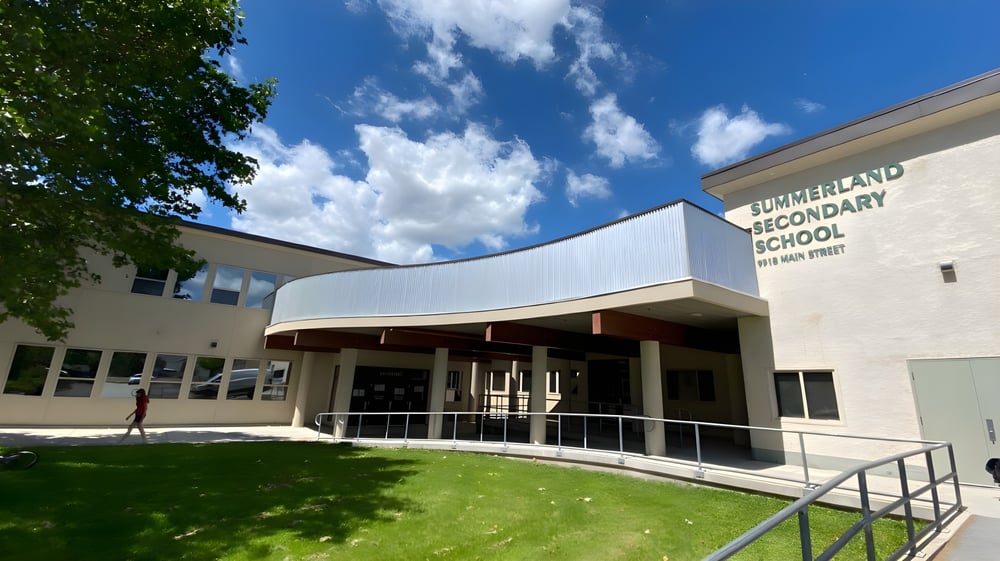 Das moderne zweigeschossige Gebäude der Summerland Secondary School mit geschwungenem Eingang und Rasen im Vordergrund unter blauem Himmel.