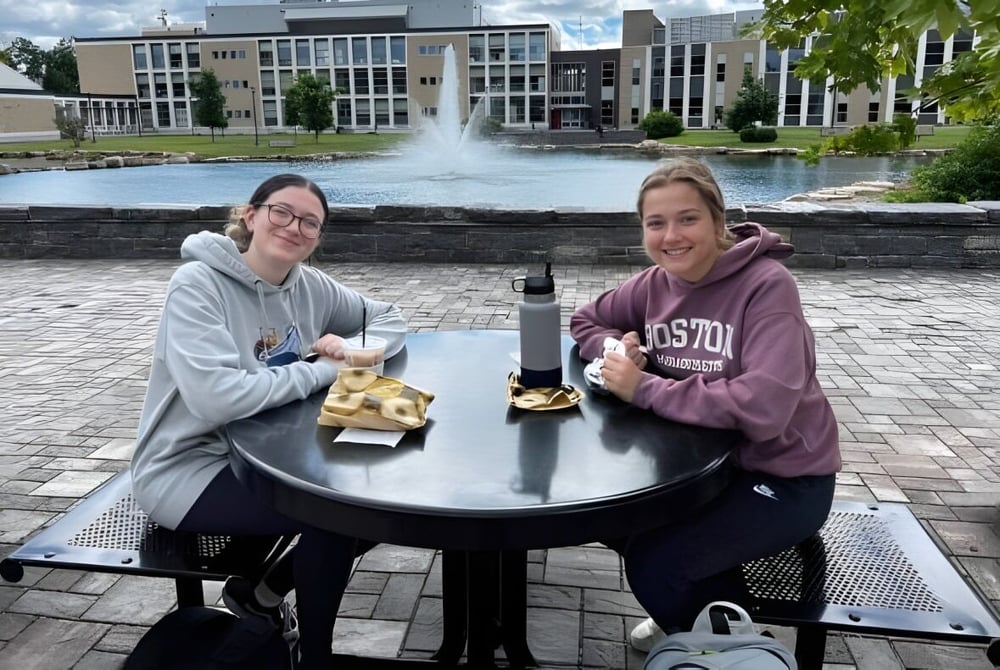 Zwei Schülerinnen der SUNY Plattsburgh sitzen an einem Tisch vor einem Brunnen und essen einen Snack auf dem Campus.