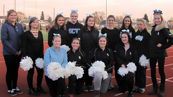 Eine Gruppe von Cheerleadern steht auf dem Sportfeld des Superior Collegiate & Vocational Institute mit weißen Pompons in schwarzen Uniformen.