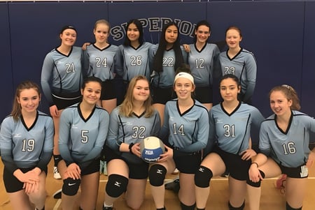 Eine Gruppe junger Volleyballspielerinnen in grauen Uniformen posiert in der Turnhalle des Superior Collegiate & Vocational Institute.