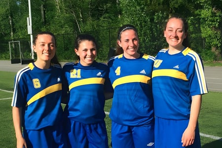 Vier Schülerinnen der Sutherland Secondary School stehen gemeinsam in blauen und gelben Sportuniformen auf einem grasbewachsenen Spielfeld.