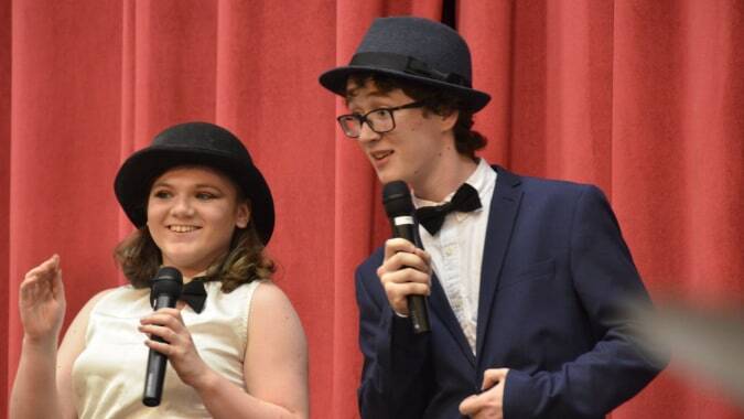 Eine Frau und ein Mann stehen auf der Bühne der Sutton Park School und sprechen mit Mikrofonen vor einem roten Vorhang.