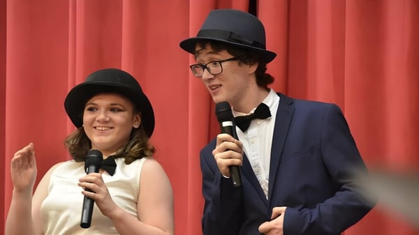 Zwei Personen stehen auf der Bühne der Sutton Park School und sprechen mit Mikrofonen vor rotem Vorhang.