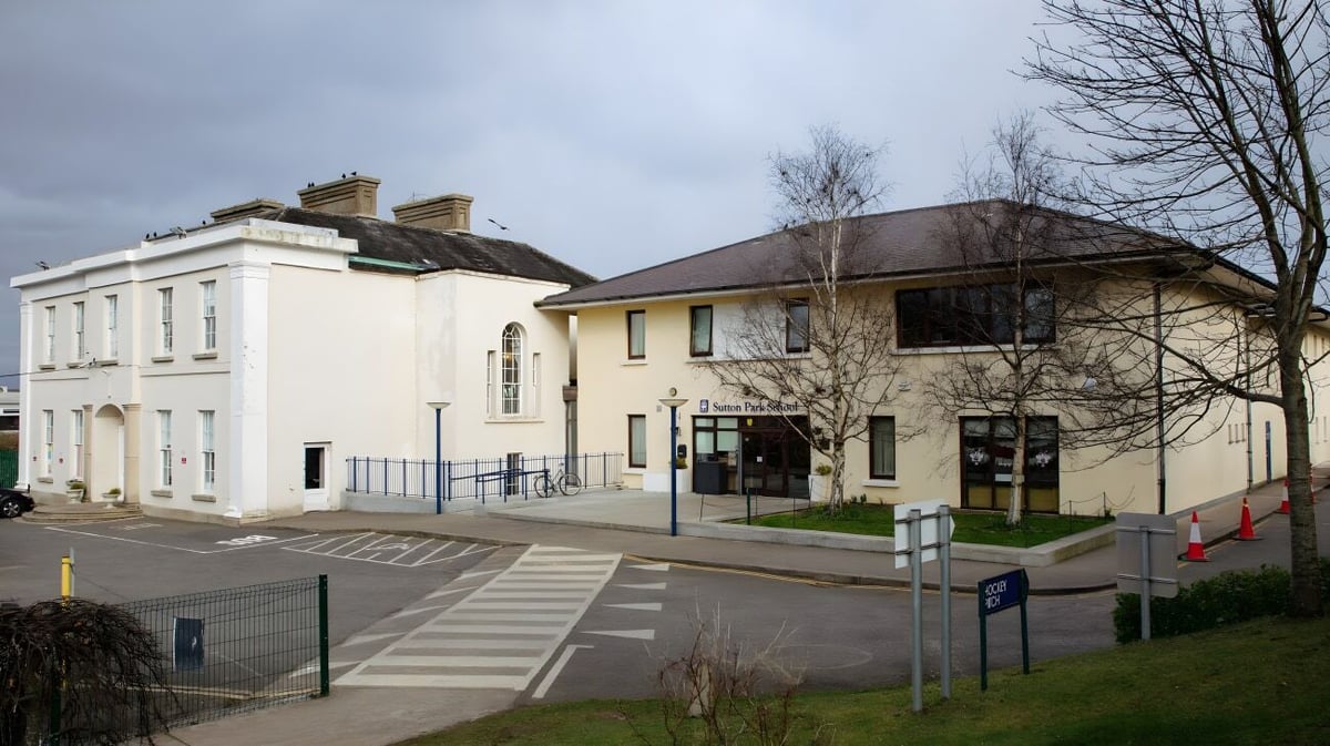 Das zwei-geschossige Schulgebäude der Sutton Park School in Dublin mit weißer Fassade und schrägem Dach ist von kahlen Bäumen umgeben.