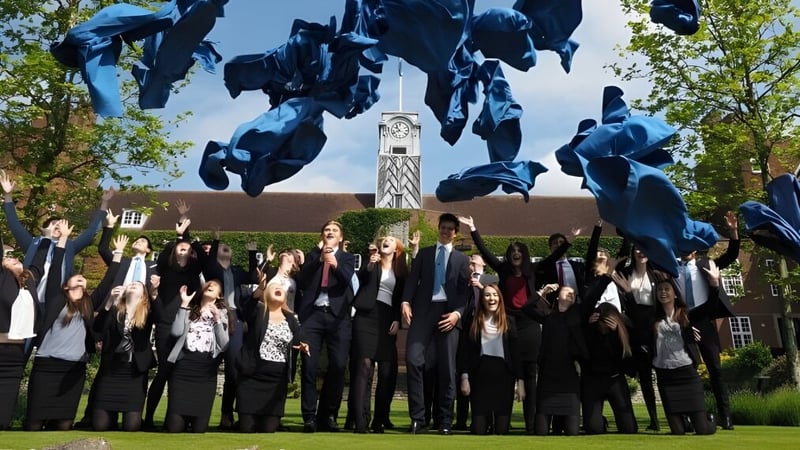 Eine Gruppe Absolventen der Sutton Valence School steht in schwarzen Talaren vor einer großen abstrakten Skulptur mit Bäumen im Hintergrund.