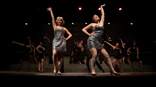 Zwei Frauen in silbernen Kleidern stehen auf der Bühne bei einem Auftritt der Tabor Academy mit tanzenden Schülerinnen und Schülern im Hintergrund.