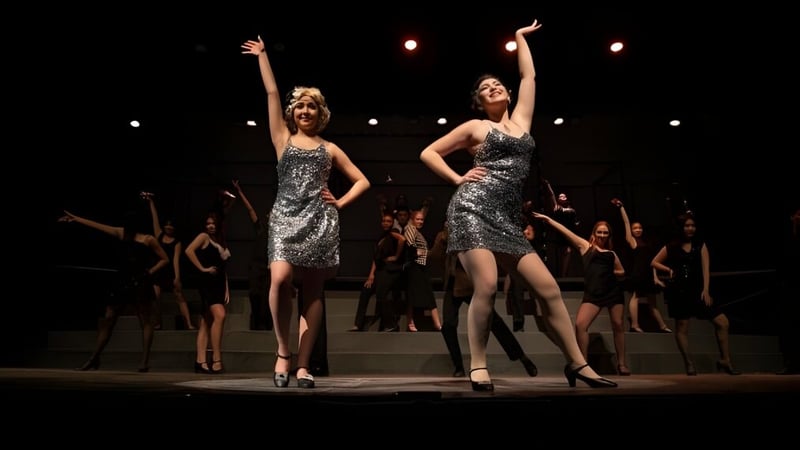 Zwei Frauen in silbernen Kleidern stehen auf der Bühne bei einem Auftritt der Tabor Academy mit tanzenden Schülerinnen und Schülern im Hintergrund.