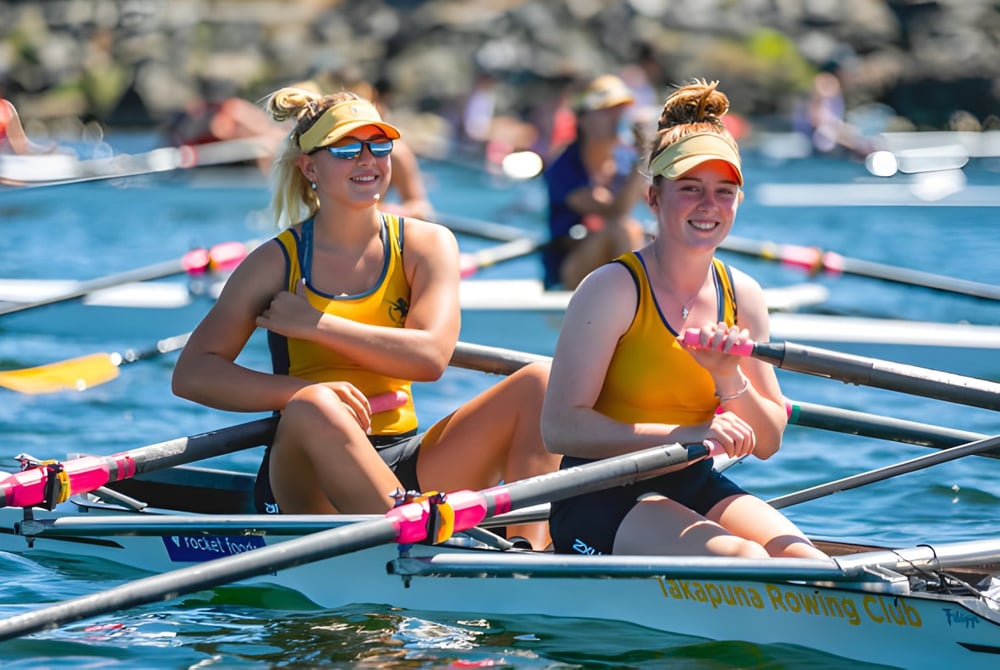 Zwei Schülerinnen der Takapuna Grammar School sitzen in einem kleinen Ruderboot auf dem Wasser vor Zuschauern.