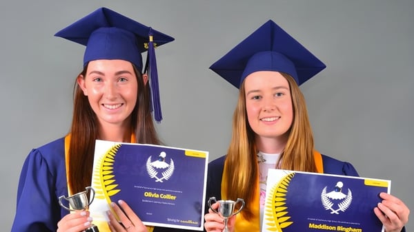 Zwei Schülerinnen der Taumarunui High School tragen Abschlusskleidung und halten ihre Diplome vor grauem Hintergrund.