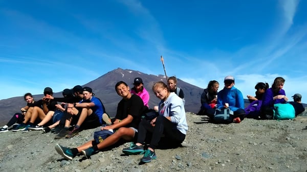 Eine Gruppe von Schülerinnen und Schülern der Taumarunui High School sitzt vor einem schneebedeckten Berg unter blauem Himmel.
