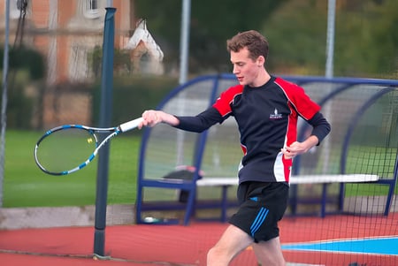 Ein Schüler der Taunton School International spielt Tennis auf einem bunten Außenplatz mit Gebäuden und Bäumen im Hintergrund.