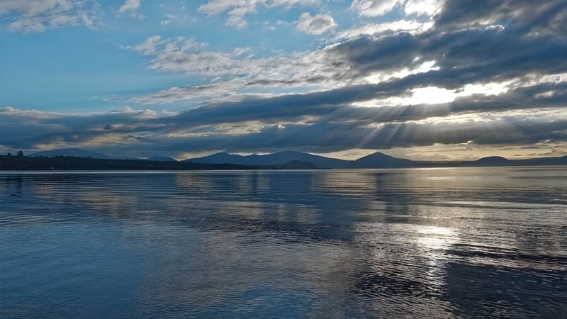 Ein ruhiger See mit reflektierendem Himmel und Sonnenstrahlen vor den Bergen, nahe dem Taupo-nui-a-Tia College.