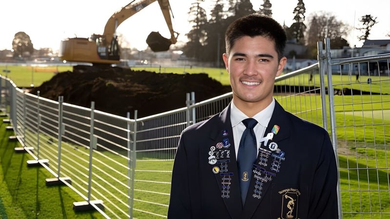 Ein Schüler von Tauranga Boy's College steht lächelnd in Uniform vor einem Sportplatz mit Baugeräten im Hintergrund.