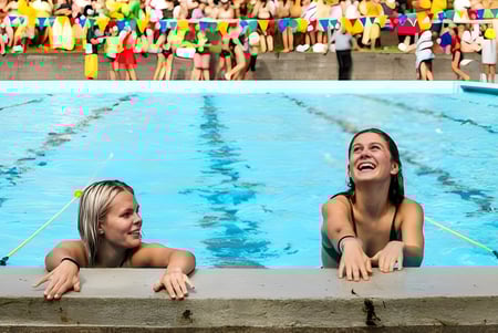 Zwei Frauen sitzen im Schwimmbecken auf dem Gelände des Tauranga Girl´s College, während im Hintergrund eine Menschenmenge und bunte Fahnen zu sehen sind.