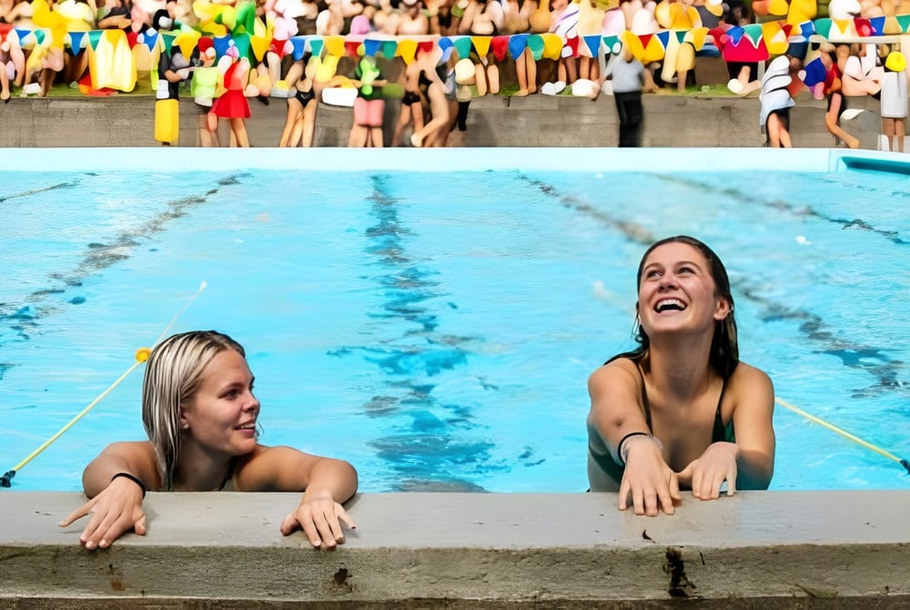 Zwei Frauen sitzen im Schwimmbecken auf dem Gelände des Tauranga Girl´s College, während im Hintergrund eine Menschenmenge und bunte Fahnen zu sehen sind.