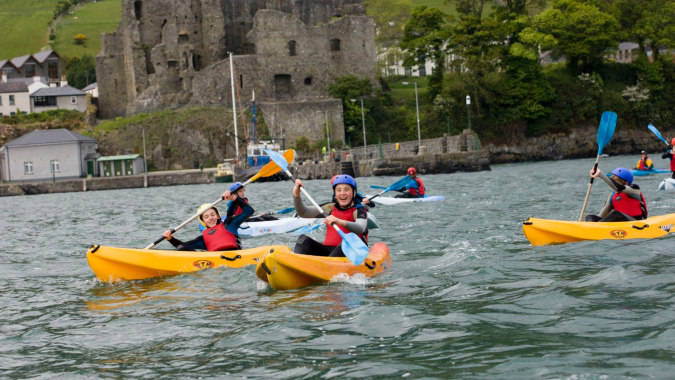 Eine Gruppe von Schülern vom Templemichael College paddelt im Kajak vor einer historischen Burg inmitten grüner Natur.
