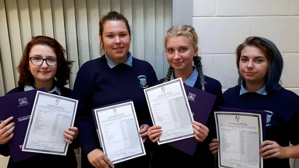 Vier Schülerinnen in Schuluniform halten Urkunden vor einer Wand mit vertikalen Jalousien auf dem Gelände des Templemichael College.