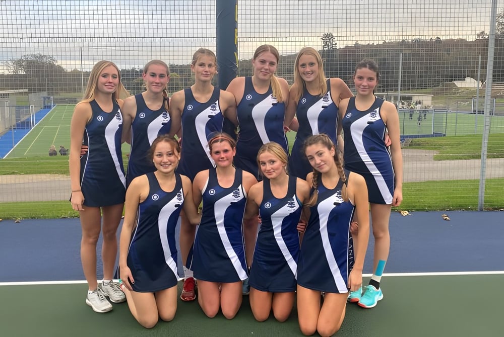 Eine Gruppe junger Sportlerinnen steht gemeinsam auf dem Sportfeld der Templemichael College mit Zaun und Bäumen im Hintergrund.