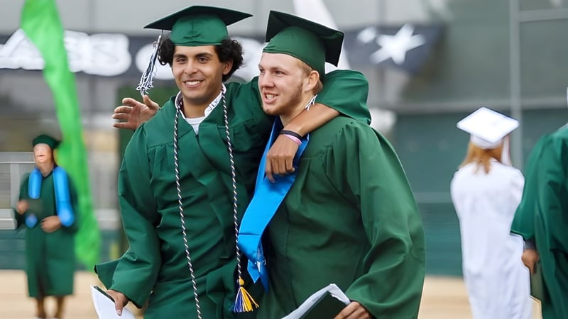 Zwei Absolventen der Templeton Secondary School umarmen sich in grünen Talaren vor weiteren Absolventen.