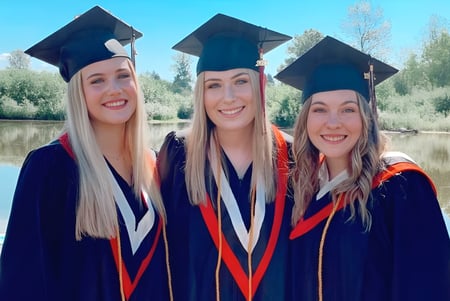 Drei Schülerinnen der Terry Fox Secondary School posieren in Abschlusstracht vor einem See mit Wald im Hintergrund.