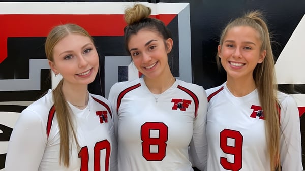 Drei Schülerinnen der Terry Fox Secondary School tragen weiße Sporttrikots mit roten Zahlen vor einem rot-schwarzen Hintergrund.