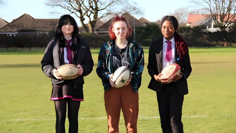 Drei Schülerinnen der The Bourne Academy stehen auf einem Grasfeld und halten Rugbybälle in der Hand.