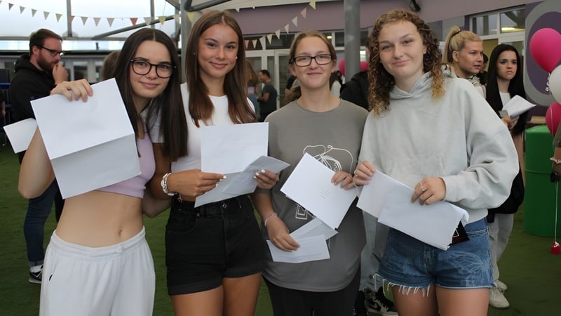 Eine Gruppe Schüler der The Bourne Academy hält Papiere und steht vor einem Gebäude mit bunten Fahnen.