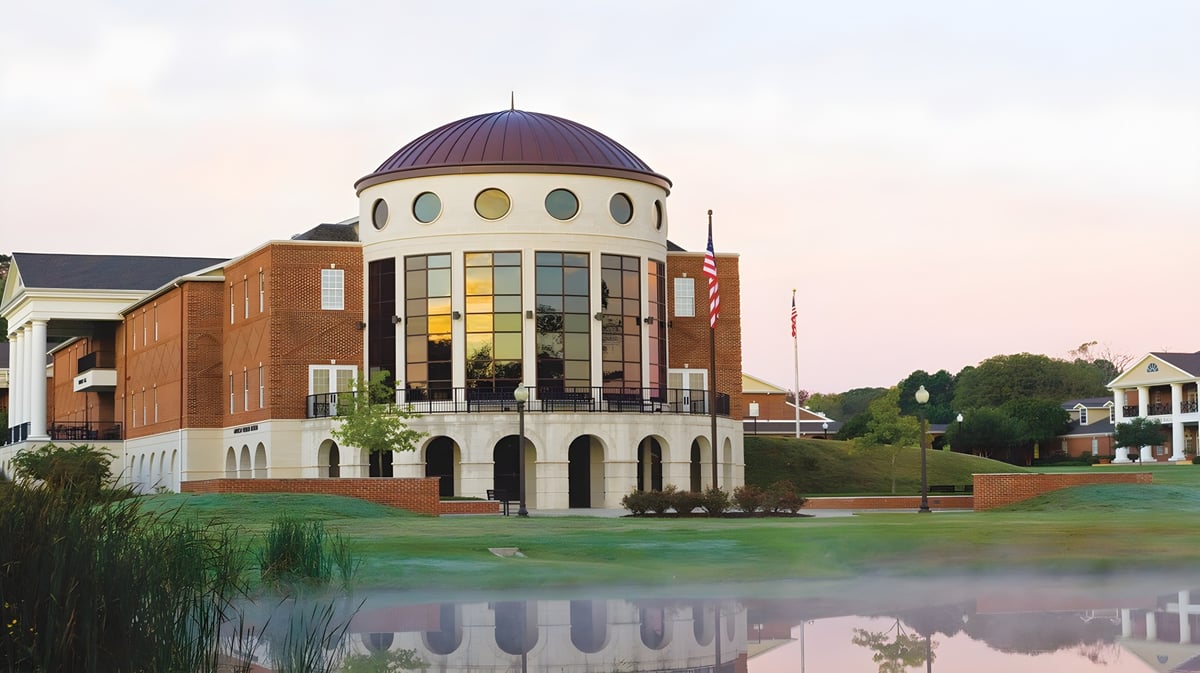 Das mehrstöckige Kuppelgebäude der The Brook Hill School spiegelt sich im ruhigen Wasser eines Teichs vor einem grasbewachsenen Hügel.