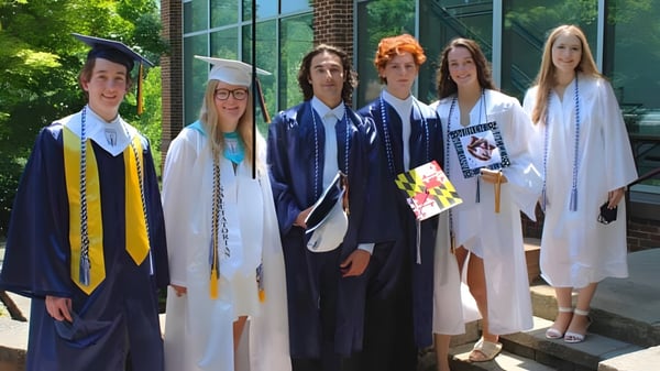 Eine Gruppe von Absolventinnen und Absolventen der The Calverton School steht in akademischer Kleidung vor einem Backsteingebäude mit großen Fenstern und Grünpflanzen.