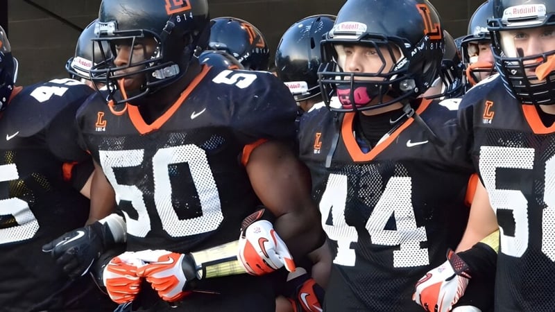 Eine Gruppe Football-Spieler der The Linsly School steht in schwarzen und orangefarbenen Uniformen mit Helmen auf dem Spielfeld.