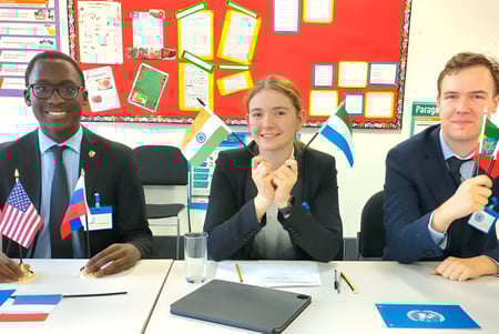 Drei Schüler sitzen an einem Unterrichtstisch im Klassenraum des National Mathematics and Science College vor einer Pinnwand mit internationalen Flaggen.