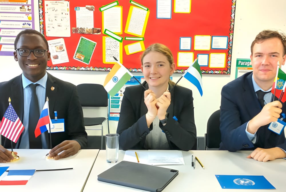 Drei Schüler sitzen an einem Unterrichtstisch im Klassenraum des National Mathematics and Science College vor einer Pinnwand mit internationalen Flaggen.