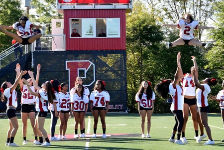 Schüler der The Pennington School führen akrobatische Cheerleader-Stunts auf einem Sportfeld vor einem roten und weißen Gebäude aus.