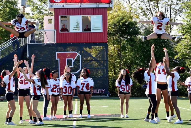 Schüler der The Pennington School führen akrobatische Cheerleader-Stunts auf einem Sportfeld vor einem roten und weißen Gebäude aus.