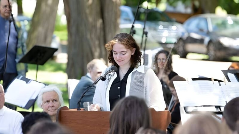 Eine Frau mit lockigen Haaren hält eine Rede am Rednerpult vor einer Gruppe von Menschen auf dem Campus der The Putney School.