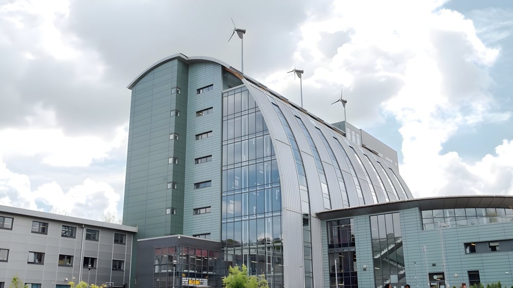 Das moderne Gebäude des The Sheffield College mit Glasfassade und Windkraftanlagen auf dem Dach unter bewölktem Himmel.