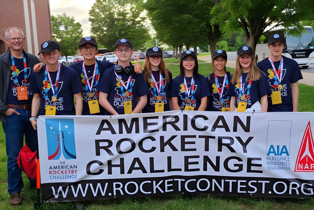 Schüler der The Vanguard School posieren vor einem Banner der American Rocketry Challenge mit Bäumen und Gebäuden im Hintergrund.