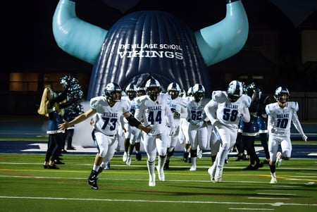 Schüler der The Village School laufen in weißen Trikots auf das Spielfeld vor einem großen aufblasbaren Wikingerhelm.