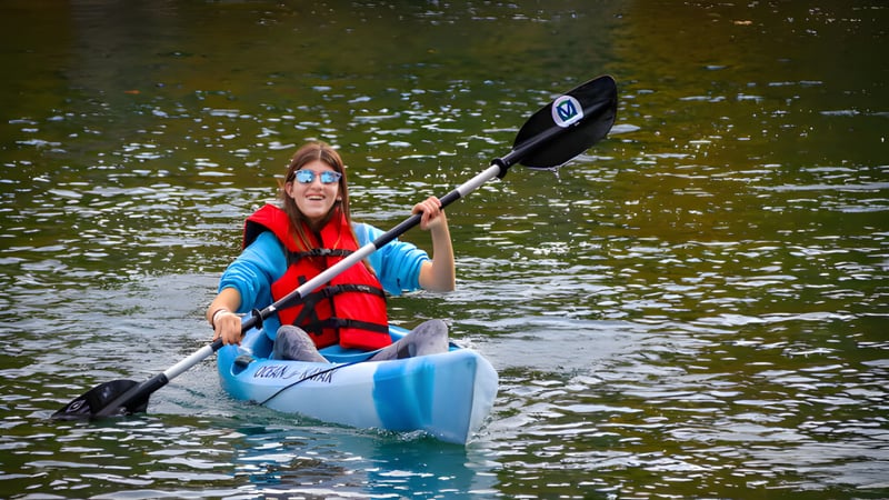 Eine Person in roter Schwimmweste paddelt mit einem blauen Kajak auf dem Wasser auf dem Gelände der The Village School.