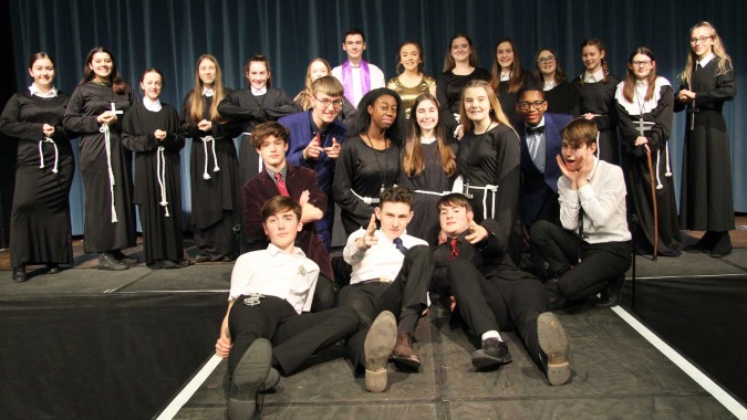 Eine Gruppe junger Menschen posiert in schwarzer Kleidung für ein Gruppenfoto auf einer Bühne der Theale Green School.