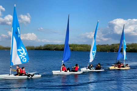 Schueler der Theale Green School segeln mit kleinen Segelbooten auf einem ruhigen See umgeben von gruener Landschaft.