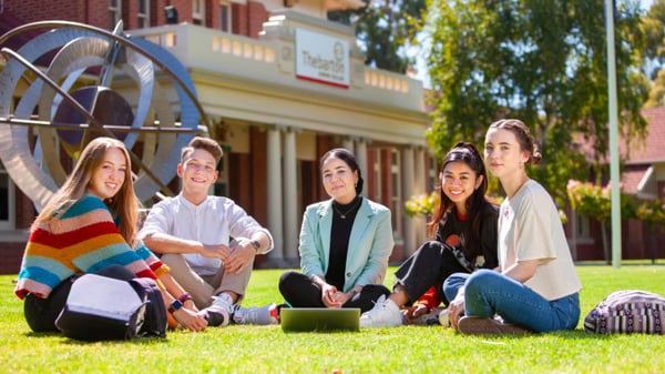 Eine Gruppe Schüler sitzt auf der Wiese vor einem Backsteingebäude mit einem großen Rad auf dem Gelände des Thebarton Senior College.