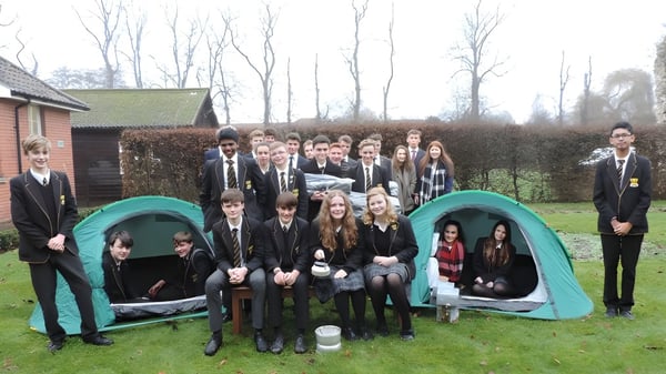 Eine Gruppe Schülerinnen und Schüler der Thetford Grammar School posiert im Freien vor einer Holzhütte und Bäumen.
