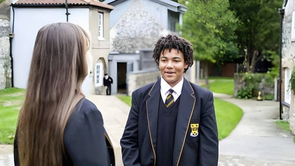 Zwei Schülerinnen und Schüler gehen gemeinsam auf einem Weg mit Grünflächen und einem Gebäude im Hintergrund auf dem Campus der Thetford Grammar School.