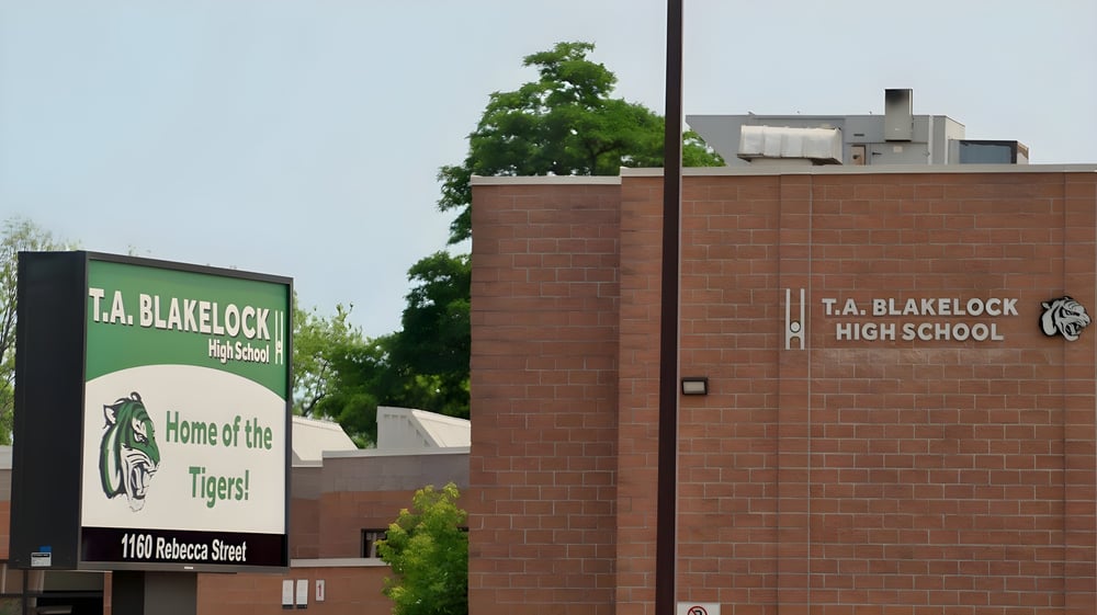 Das Backsteingebäude der Thomas A. Blakelock High School mit einem Schild 'Home of the Tigers' im Vordergrund ist von Bäumen umgeben.